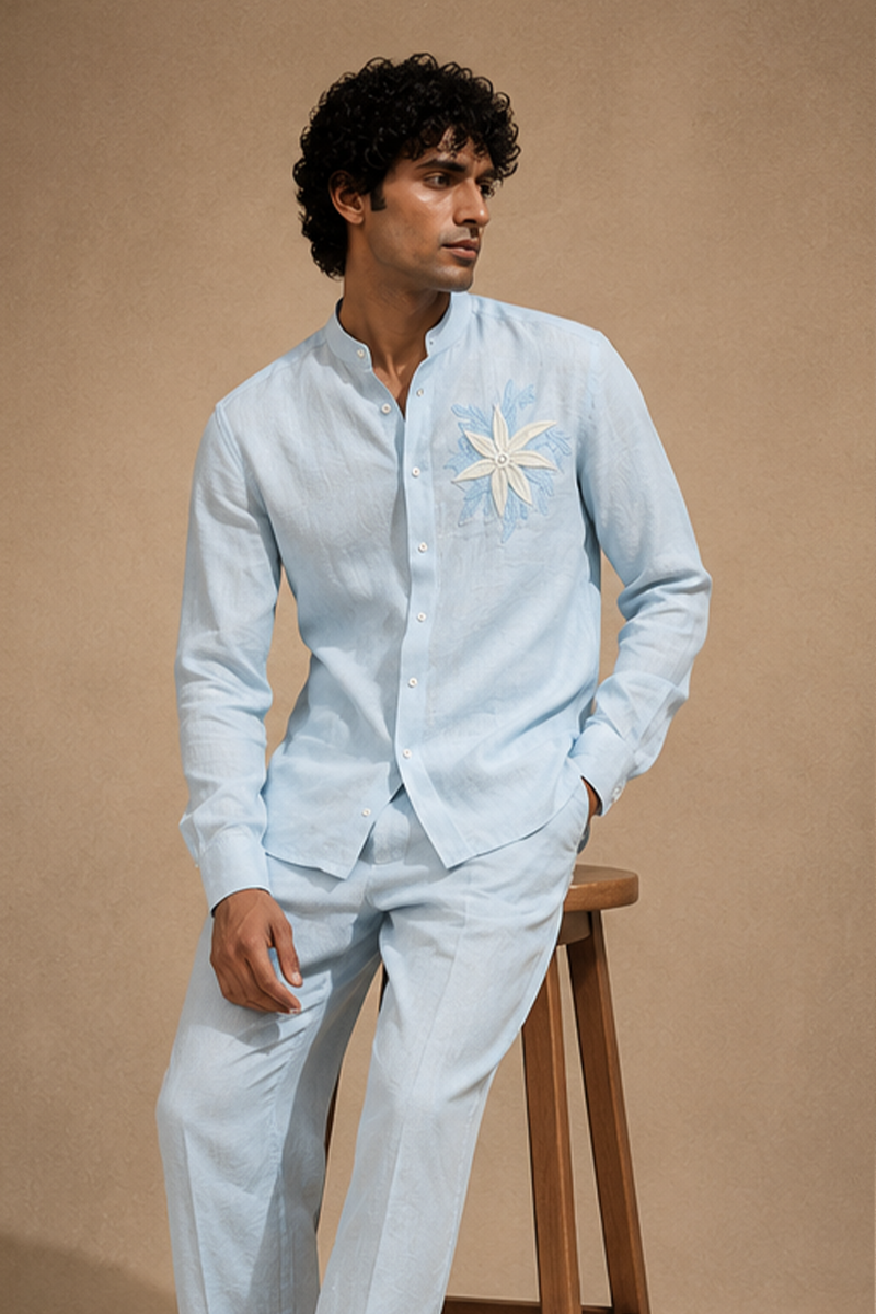 Male model seated on a wooden stool wearing a sky-blue linen co-ord set with a mandarin collar shirt, matching trousers, and Coral Water Flower-inspired chest embroidery.