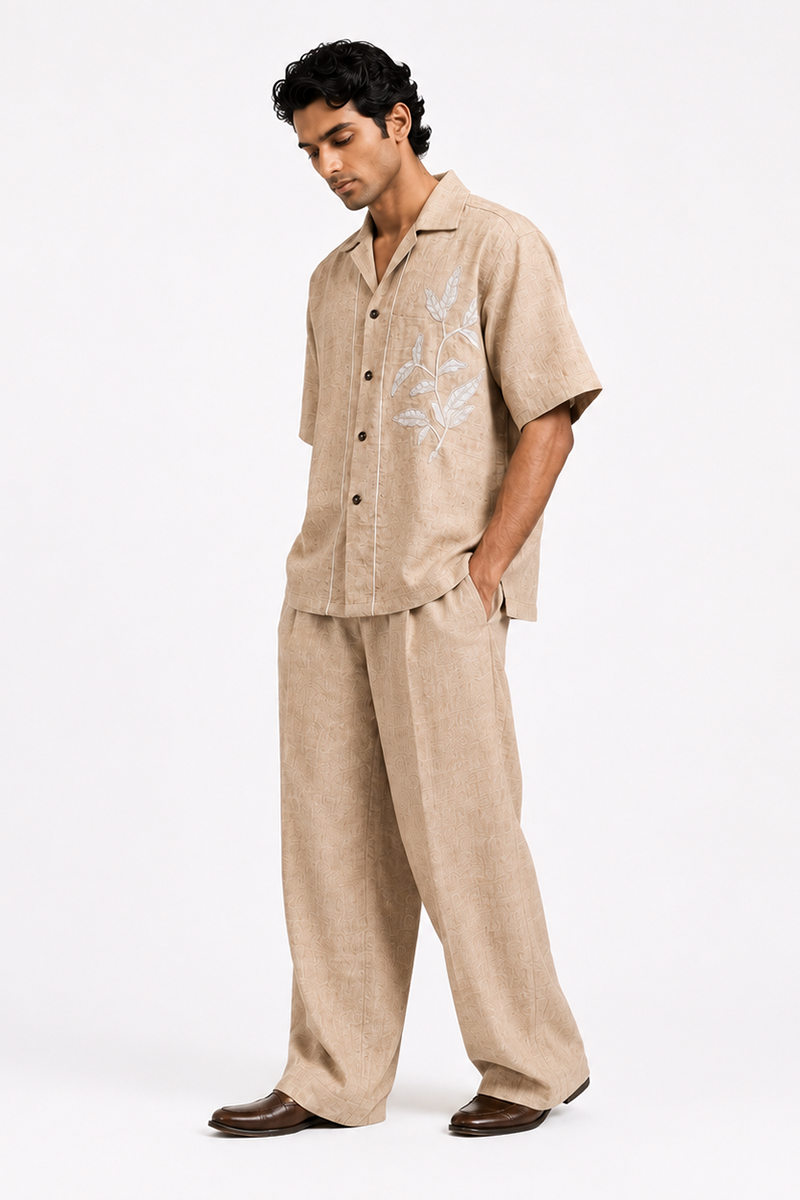 Male model wearing a beige Kalpalata-inspired cotton suiting coord set with a relaxed short-sleeve shirt, wide-leg trousers, subtle textured motif, and white floral embroidery on the chest.