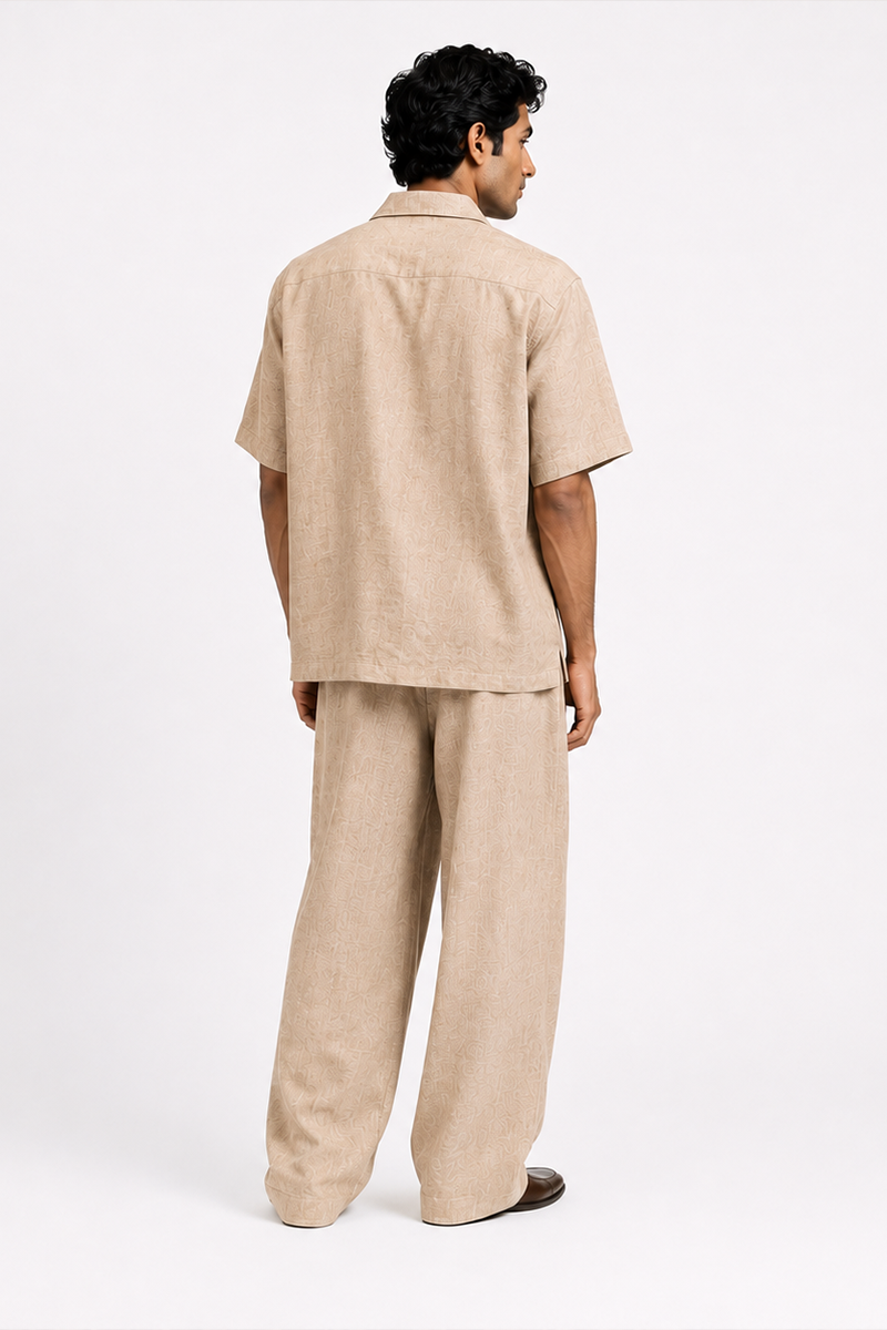 Back view of a male model wearing a beige Kalpalata-inspired cotton suiting coord set with a relaxed short-sleeve shirt and wide-leg trousers featuring a subtle textured motif.