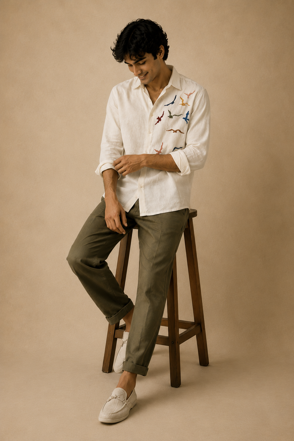 Male model seated on a wooden stool wearing a white linen shirt with colourful bird embroidery, olive trousers, and white loafers against a beige textured background.