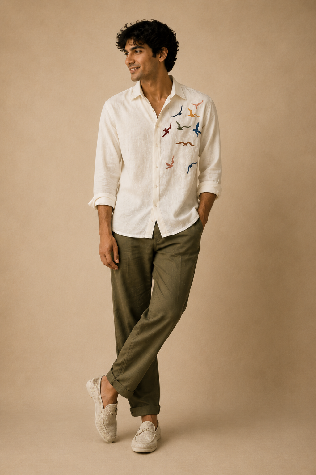 Male model wearing a white linen shirt with colourful bird embroidery, olive trousers, and white loafers, standing against a beige textured background.