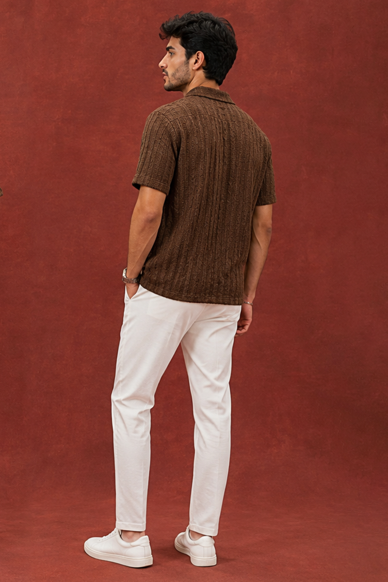 Back view of an Indian male model wearing a brown textured short-sleeve shirt with white trousers and white sneakers against a rust red backdrop.