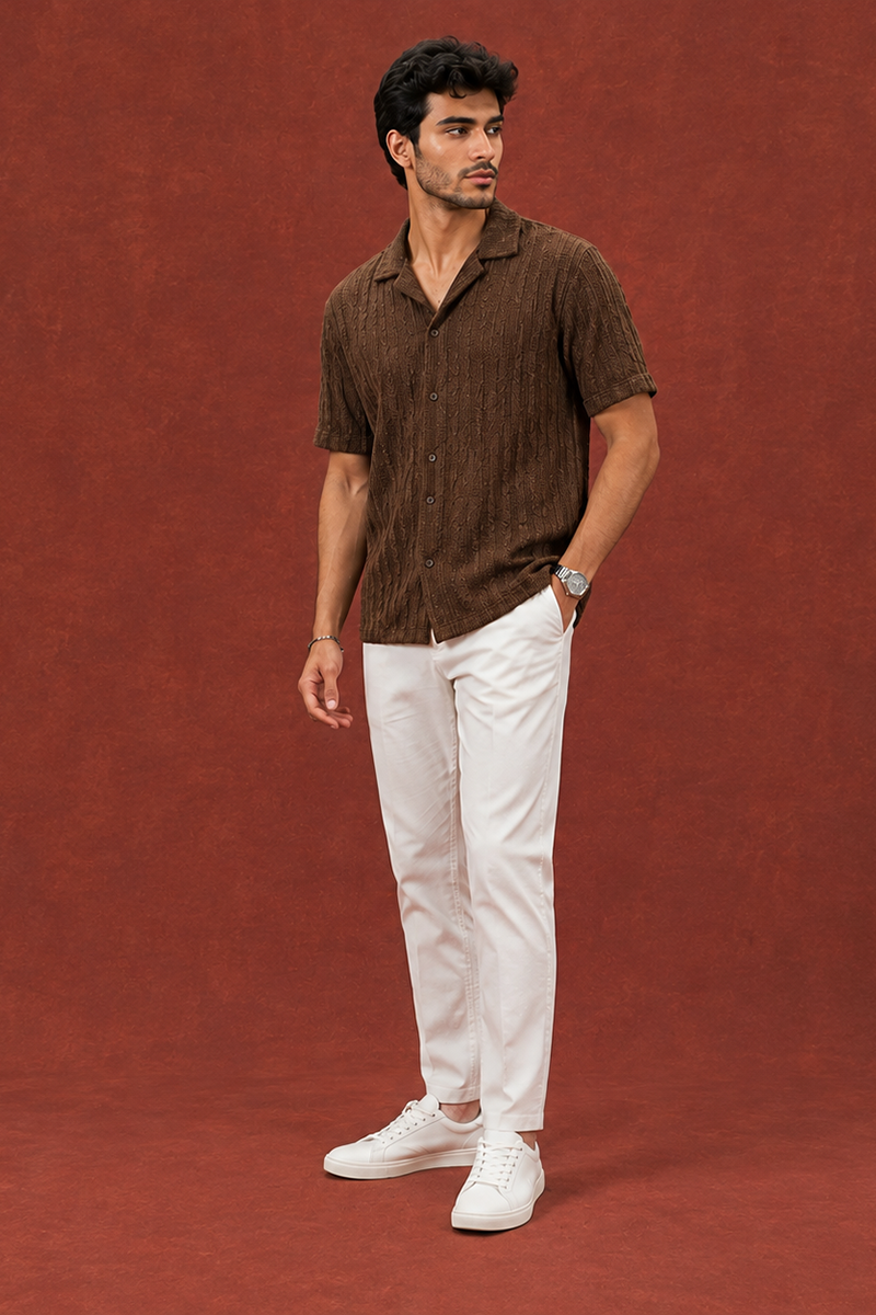 Indian male model wearing a brown textured short-sleeve shirt with white trousers and white sneakers, standing against a rust red backdrop.