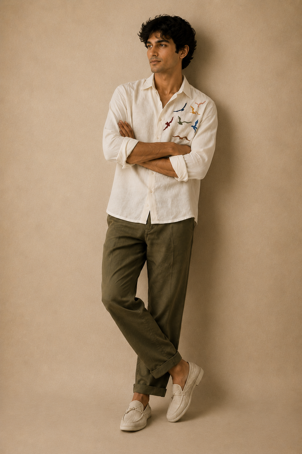 Male model wearing a white linen shirt with colourful bird embroidery on the chest, paired with olive trousers and white loafers against a beige textured background.